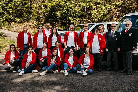 Start-01 Die Helfer:innen in weißen Polos und roten Fleecejacken sitzend und stehend als Gruppe. Alle lachen und sehen glücklich aus.