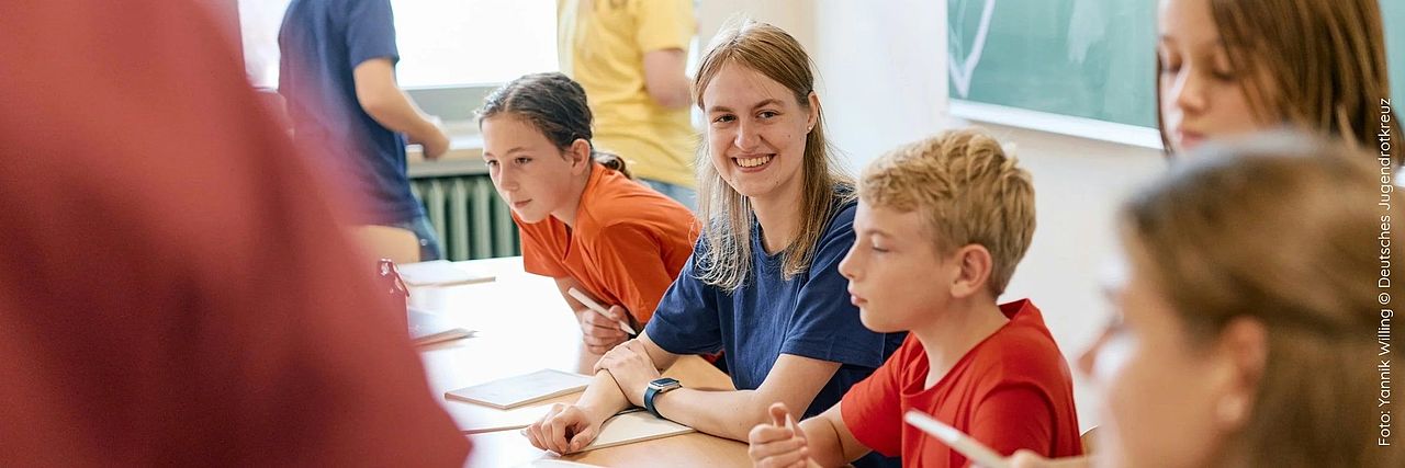 Mehrere Kinder in unterschiedlich farbigen knalligen T-Shirts sitzen an einem Tisch. Ein Mädchen in der Mitte lacht in die Kamera.